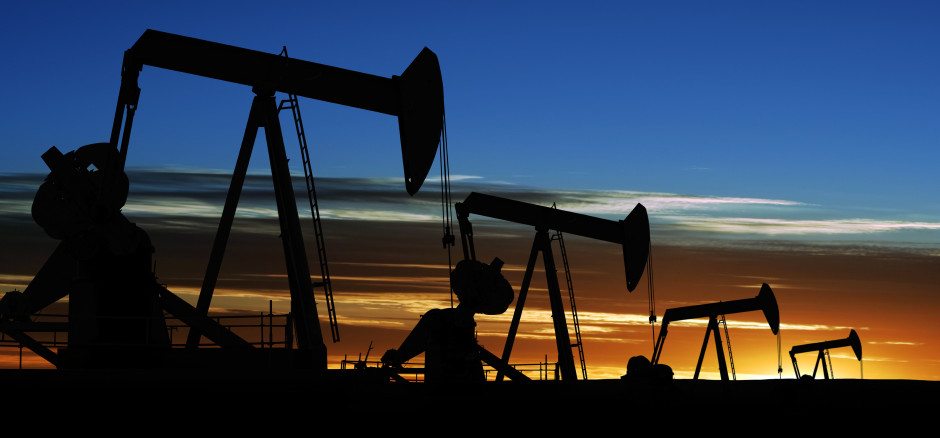 Silhouettes of several oil pumpjacks operating in a row against a dramatic sunset sky, with shades of blue, orange, and yellow on the horizon.