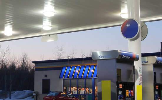 A gas station at dusk with two fuel pumps in the foreground and a convenience store with blue awnings in the background, illuminated by overhead lights.