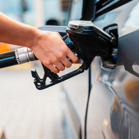 A close-up of a person’s hand holding a fuel pump nozzle, refueling a car at a gas station. The car is silver, and the focus is on the fueling process.