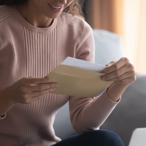 A woman sits on a couch, smiling as she opens a beige envelope and pulls out a letter. Only her upper body and hands are visible.