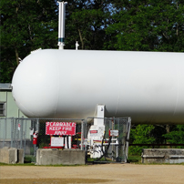 A large white propane tank sits in a fenced area outdoors, surrounded by warning signs. Lush green trees are visible in the background.