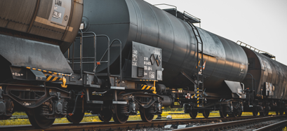Close-up view of black cylindrical tanker rail cars on train tracks, with yellow grass visible in the background under an overcast sky.