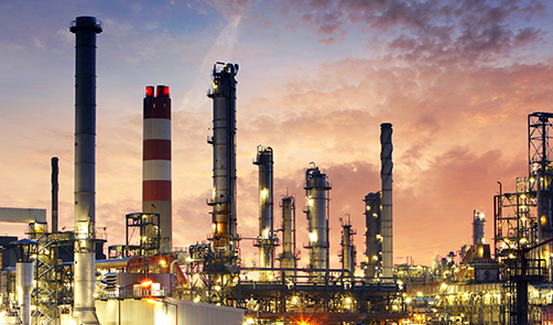 An industrial oil refinery with multiple tall chimneys and metal structures is shown at sunset, with a colorful sky in the background.