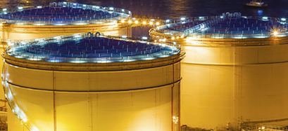 Large cylindrical storage tanks with blue tops are illuminated by bright lights at night. A body of water and a small boat are visible in the background.
