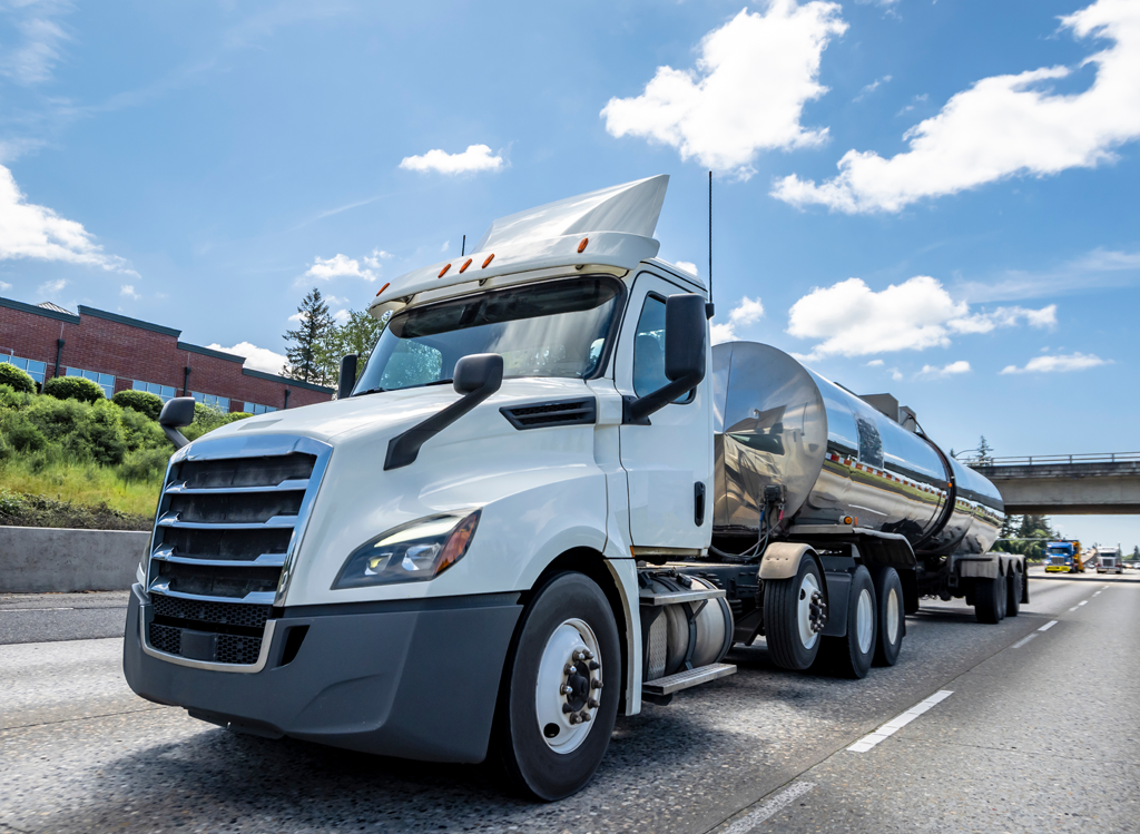 A white semi-truck with a shiny, silver tanker trailer drives down a highway under a blue sky with clouds. A bridge and a brick building are visible in the background.